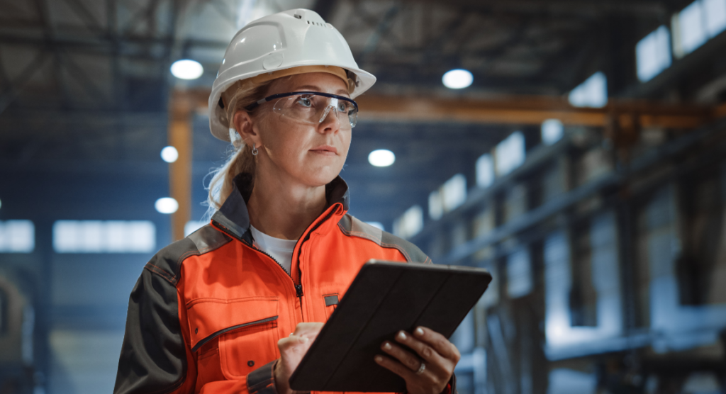 Manufacturing professional wearing a hard hat and safety glasses, using a tablet on a factory floor.